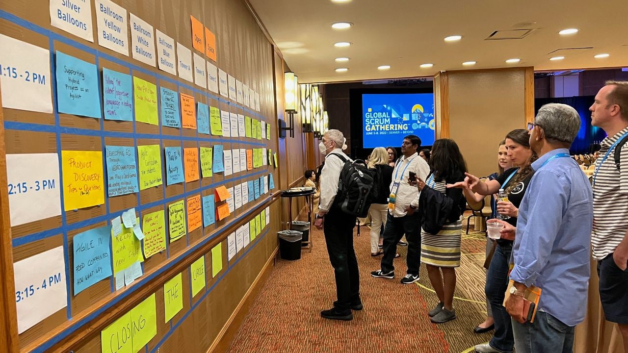 A group of Scrum Alliance Gathering attendees stand in front of a wall with a schedule of sessions listed